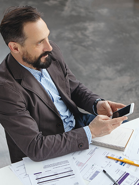 Geschäftsmann, der auf seinem Smartphone arbeitet, umgeben von Unterlagen und Notizen